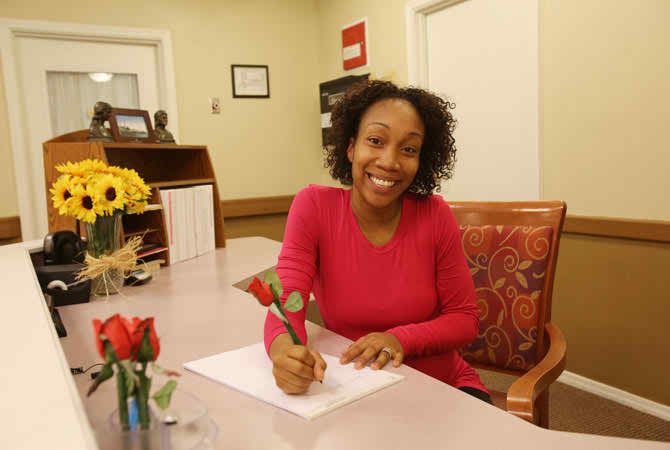 A woman smiles as she writes at a desk in an office setting, with flowers nearby.