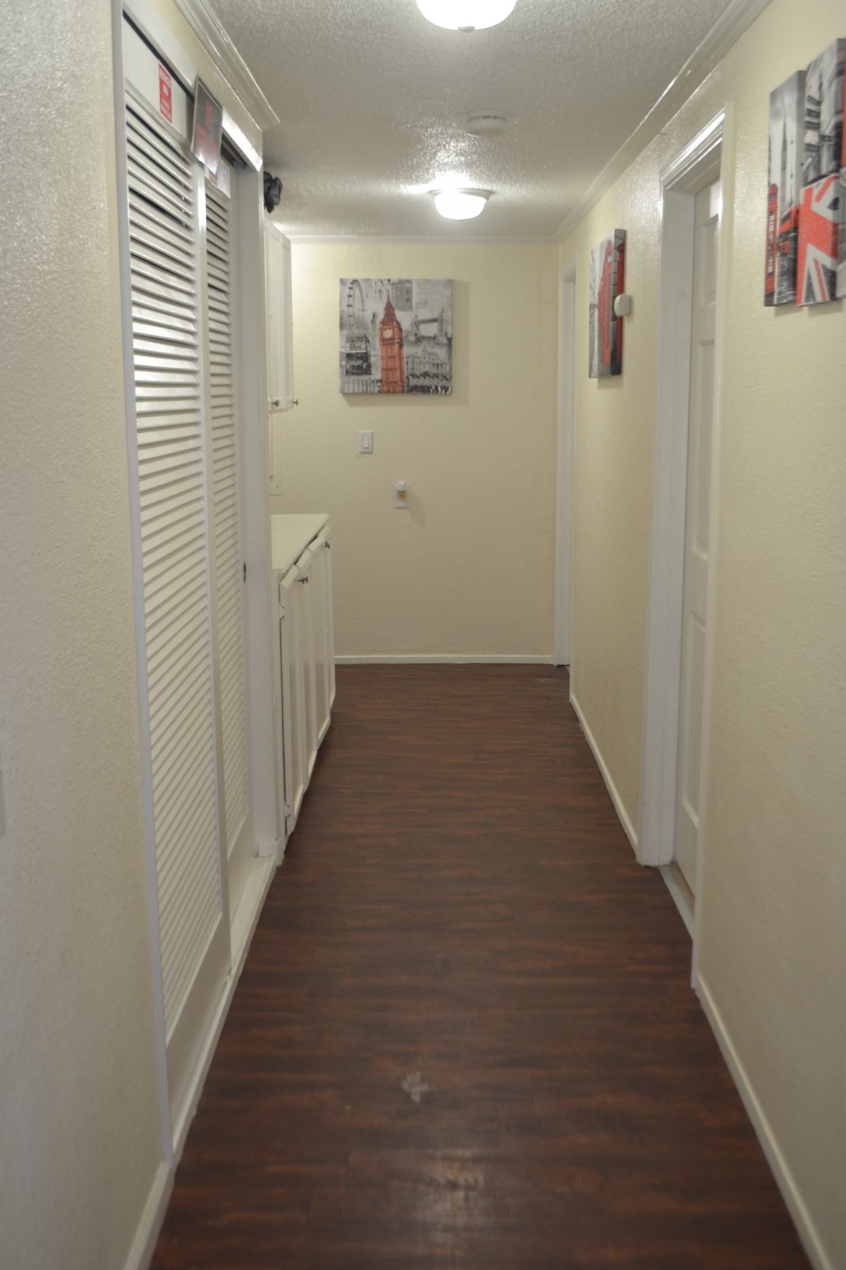 A narrow hallway with wooden flooring and white walls, decorated with paintings.