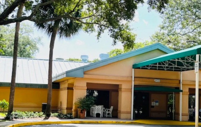 A quaint building with an entrance shaded by trees, featuring a small table and chairs outside.