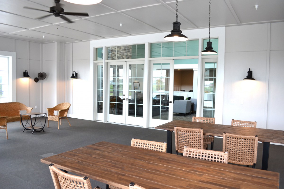 A modern indoor dining area with wooden tables and chairs, featuring pendant lights and a ceiling fan, leading to a room through glass doors.