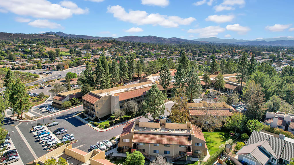 Aerial view of a residential area with multiple buildings, surrounded by trees and hills under a blue sky.