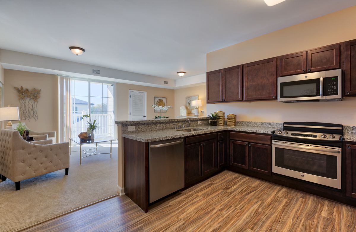 A modern kitchen with stainless steel appliances and wood flooring, connected to a bright living area.