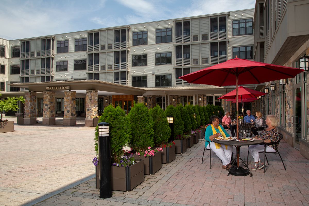 People enjoy outdoor dining on a patio with red umbrellas in an urban setting.