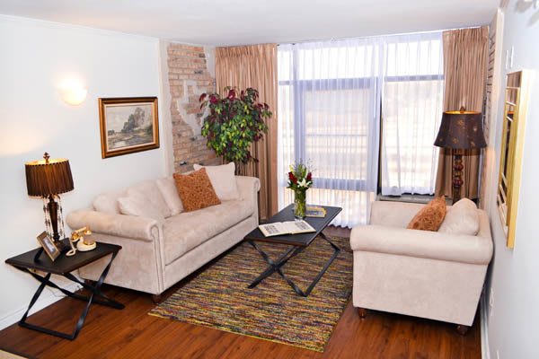 A cozy living room with beige sofas, a central coffee table, and decorative elements including an indoor plant and a painting on the brick wall.