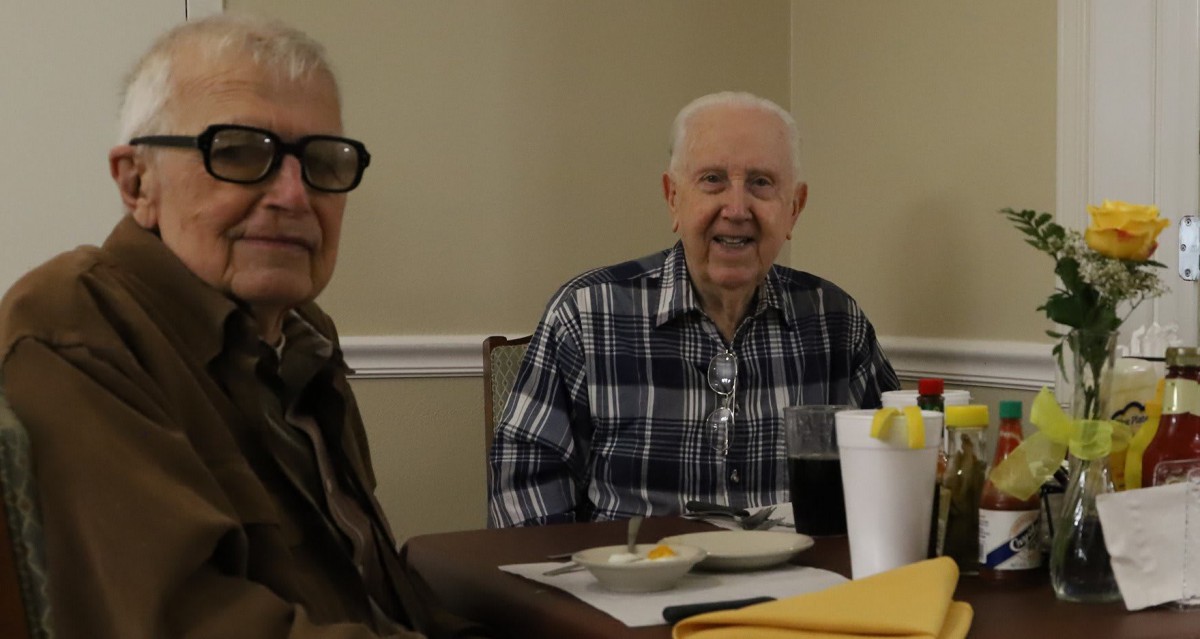 Two elderly men sitting at a dining table, one smiling.