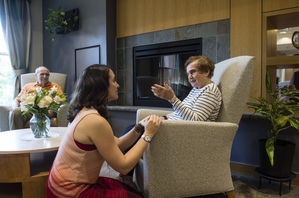 A caregiver engages in a conversation with an elderly person sitting by a fireplace.