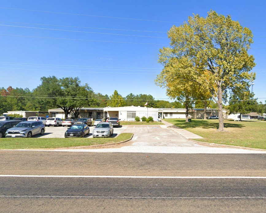 A single-story building with a parking lot and several cars, situated alongside a road on a clear day.
