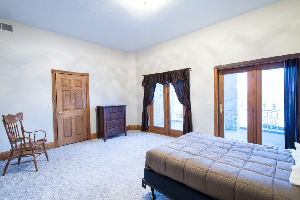 A spacious bedroom featuring a wooden dresser, large windows with curtains, and light carpet flooring.