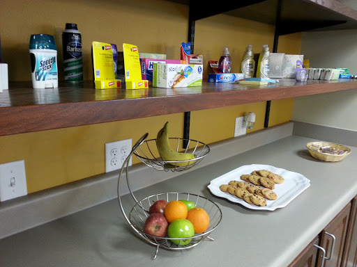 A kitchen counter displaying a fruit bowl with bananas, apples, and oranges, alongside a plate of cookies and various products on the shelf above.