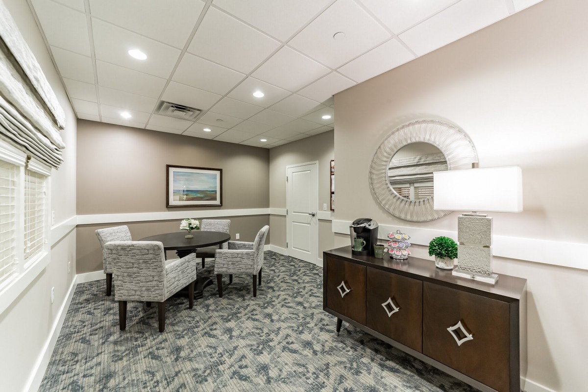 A modern office meeting room with a round table and upholstered chairs, featuring decorative elements like a mirror and lamp.