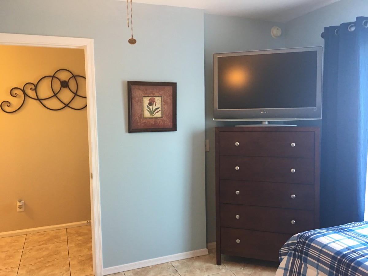 A bedroom with light blue walls featuring a wall-mounted TV above a tall dresser, adjacent to an open doorway and decorative wall art.