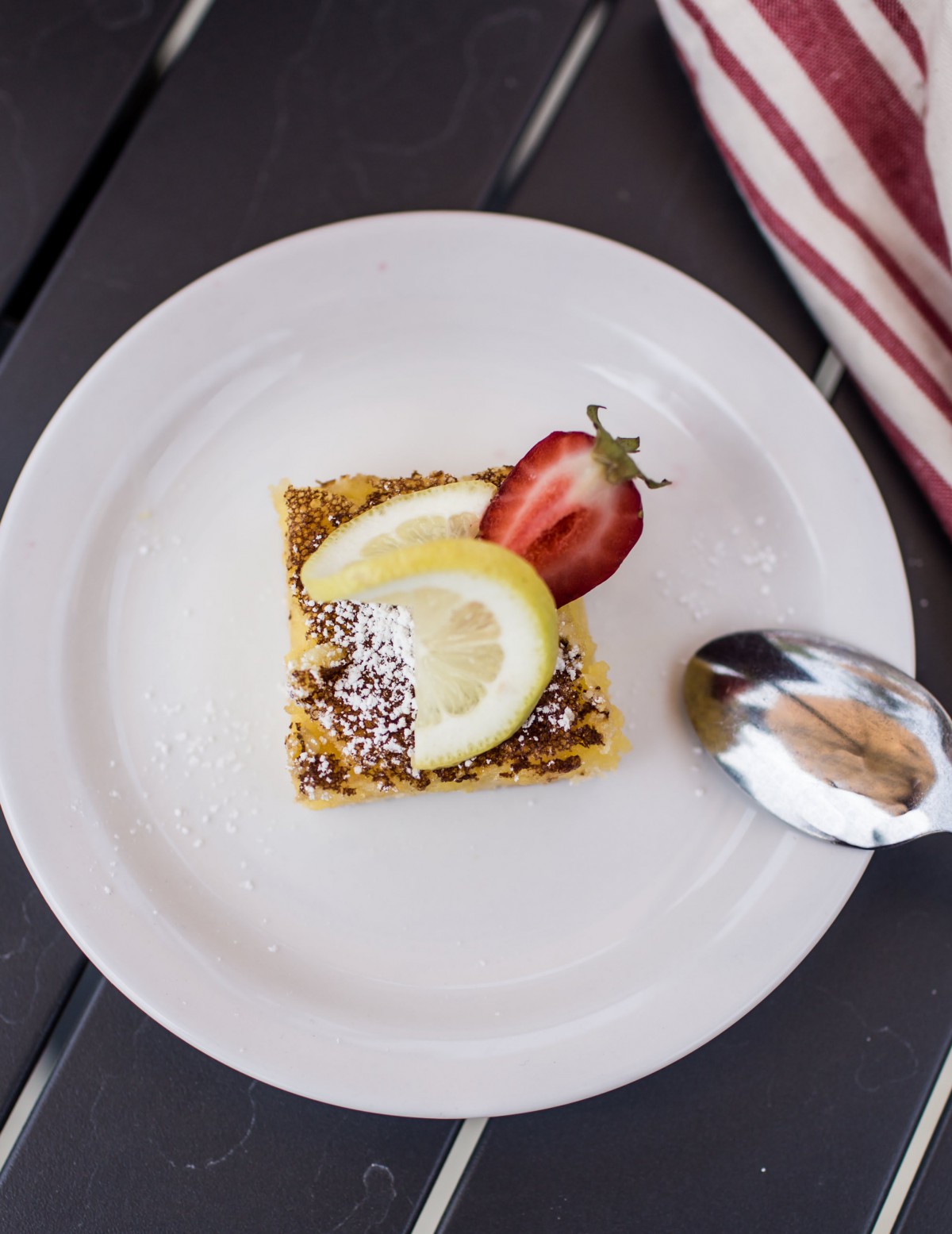 A square pastry topped with a lemon slice and strawberry, served on a white plate with a spoon.