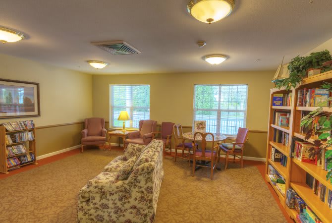 A cozy living room with a floral sofa, several chairs, and a wooden table, featuring a bookshelf and large windows.