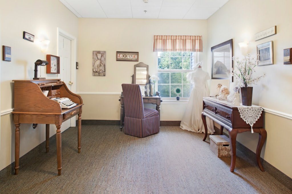 A cozy room with a vintage style, featuring a desk, chair, piano, and decorative items near a window.