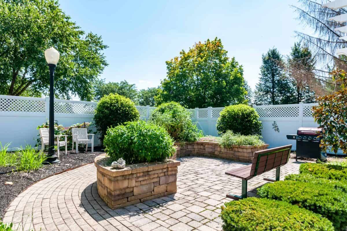 A beautifully landscaped garden with brick planters, lush greenery, and a wooden bench, enclosed by a white fence.
