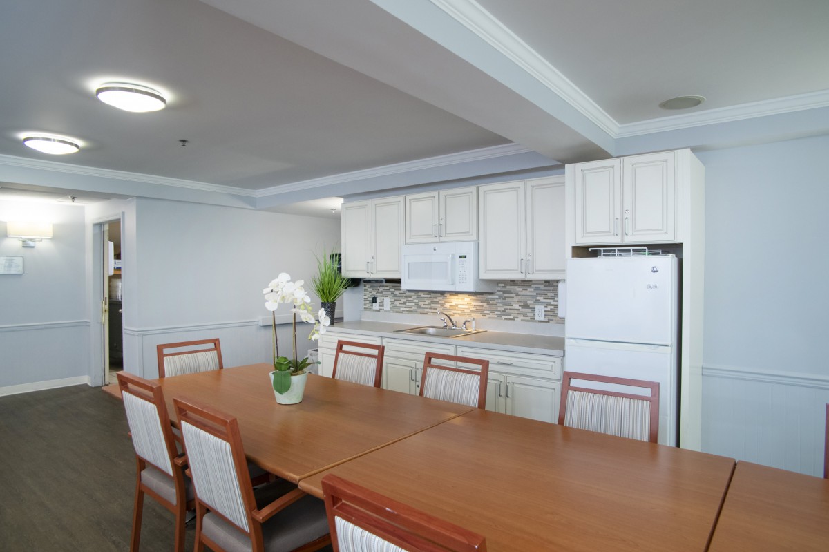 A modern dining area with a large wooden table, surrounded by chairs, adjacent to a white cabinet kitchen.