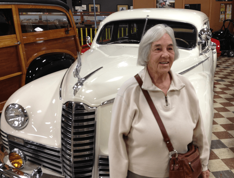 An elderly woman standing in front of a vintage car at a museum.