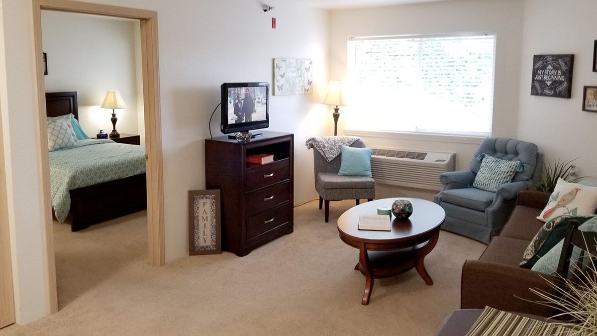 A cozy living room with modern furniture, including chairs and a round table, a television on a cabinet, and large windows allowing natural light.