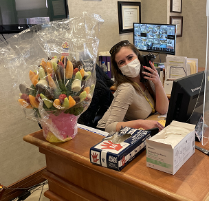 A receptionist at the desk in an office, wearing a mask and surrounded by a colorful bouquet of flowers.