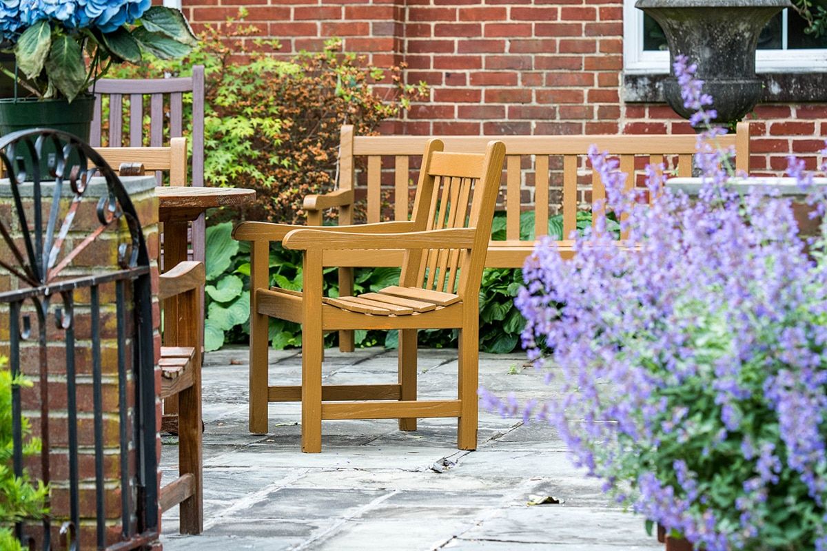 A tranquil garden patio with wooden chairs and benches surrounded by lush plants and flowers.