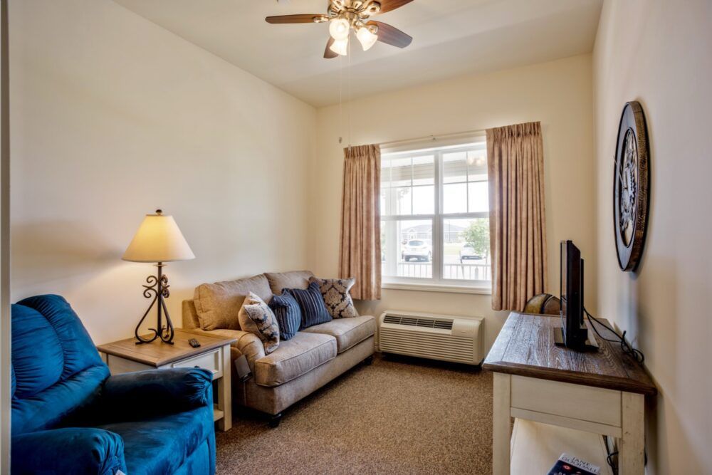 A cozy living room with a sofa, lamp, and television, featuring curtains and a ceiling fan.