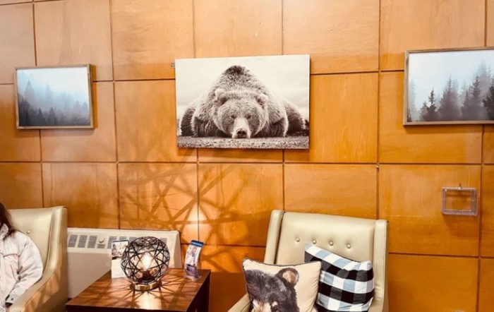 A cozy living room corner featuring a bear-themed decor with a framed bear photograph, a comfy chair adorned with pillows, and warm wood paneling.