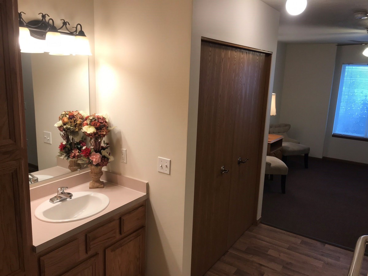 A small bathroom with a sink and mirror, leading to a hallway with visible furniture.