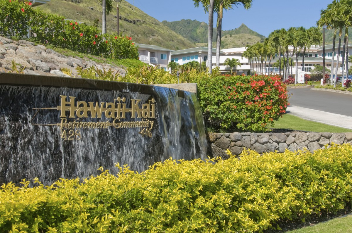 A scenic view of the Hawaii Kai retirement community entrance with lush greenery and mountains in the background.