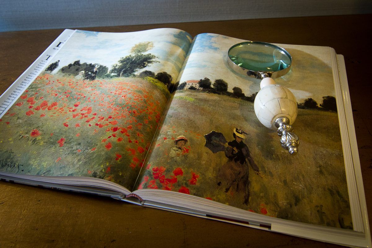An open art book displaying a landscape painting with a magnifying glass resting on top.