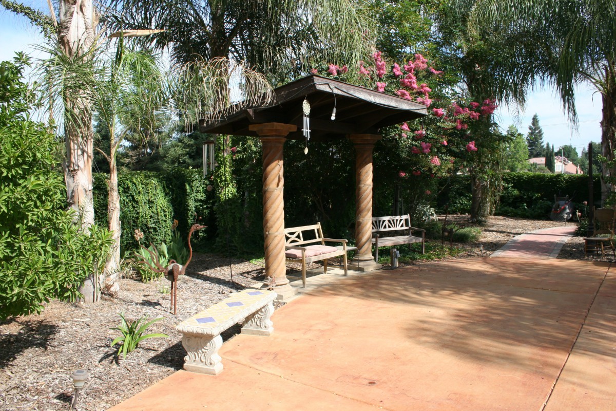A tranquil garden scene featuring a shaded pergola with benches and surrounding greenery.