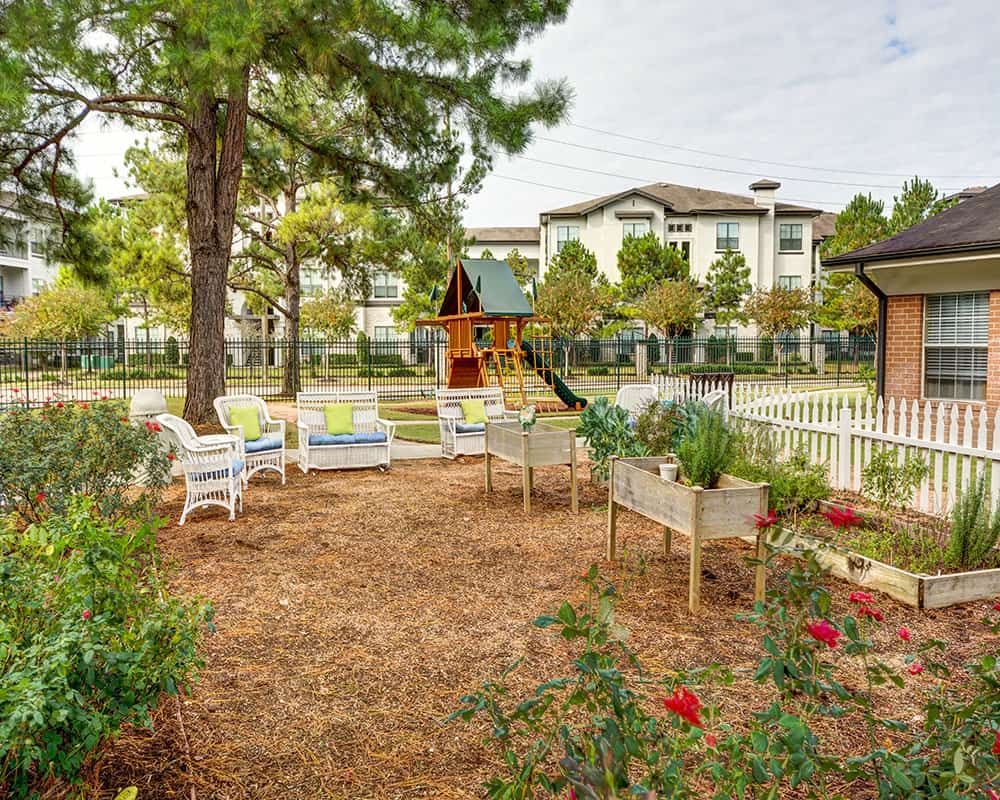 A serene garden area with chairs and a playground, surrounded by trees and overlooking a building.