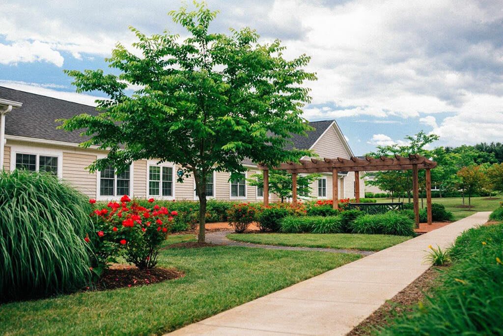 A scenic view of a house with a beautifully landscaped garden featuring trees, shrubs, and a flower bed along a sidewalk.