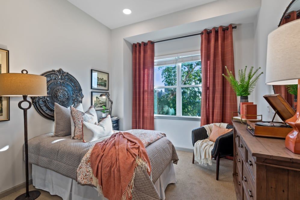 A cozy bedroom with warm-toned decor, featuring a bed with pillows and blanket, and a window with red curtains.