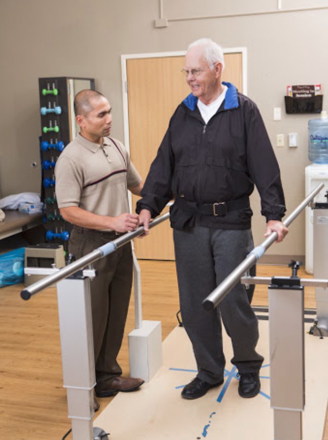 An elderly man receiving assistance from a physical therapist while walking with parallel bars in a rehabilitation center.