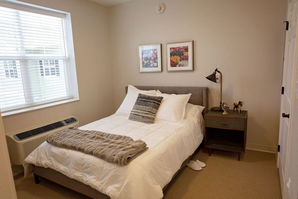 A modern bedroom with a neatly made bed, gray headboard, and decorative pillows, complemented by wall art and a nightstand with a lamp.