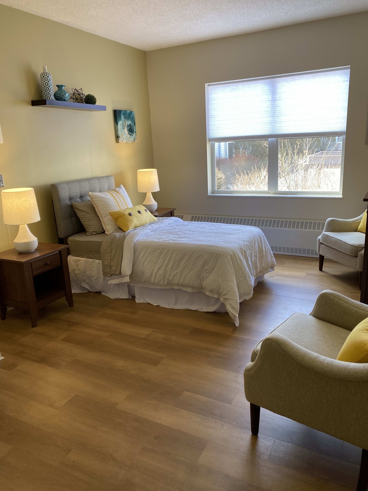 A cozy bedroom with a bed, nightstands, and chairs in a softly lit room.