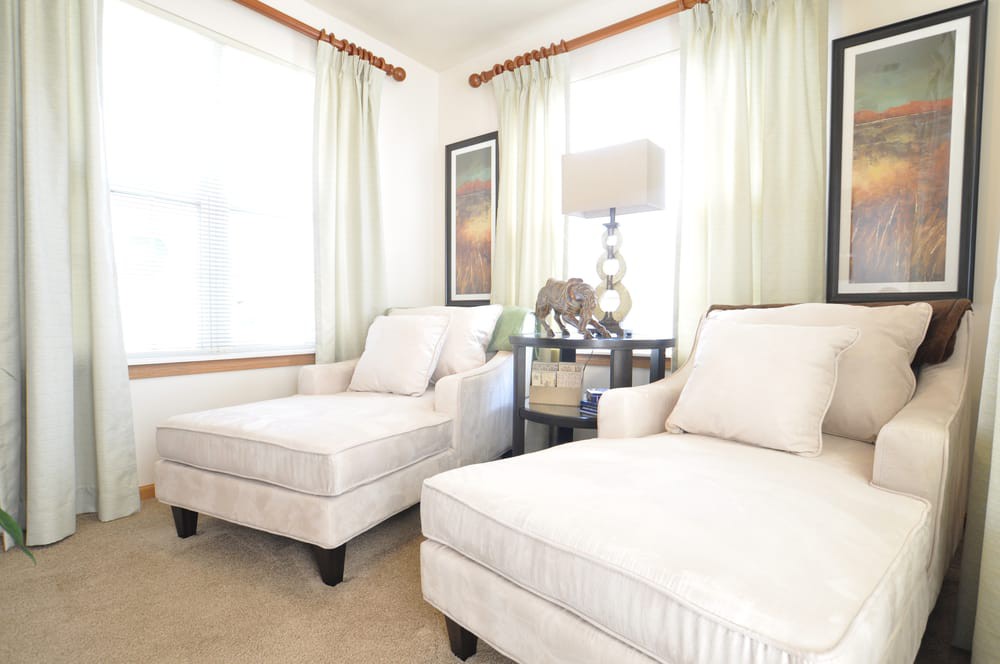 A bright and cozy living room featuring two comfortable chairs, a side table with a lamp, and large windows with light curtains.