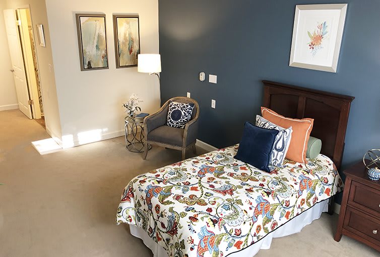 A cozy bedroom with a single bed featuring a colorful floral comforter, complemented by decorative pillows. The room is accented by wall art, a stylish chair, and a standing lamp against a navy blue accent wall.