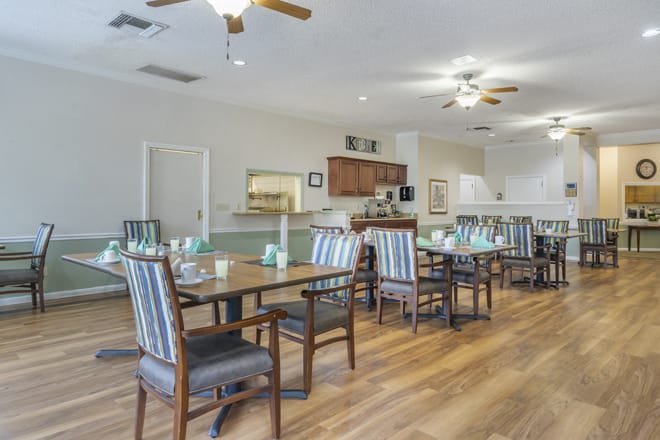 A spacious dining room with wooden floors and striped chairs, featuring a kitchen area in the background.