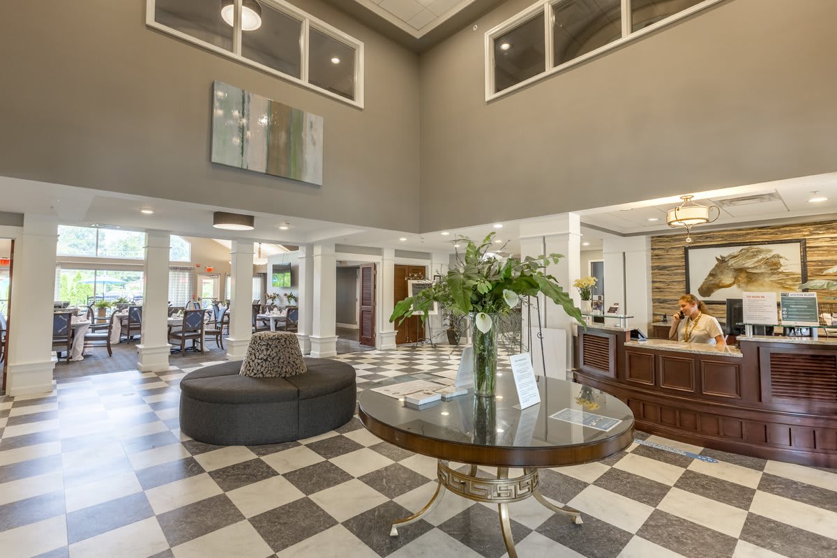 A spacious lobby area with a checkered floor, featuring a central round table adorned with flowers and surrounded by various pieces of furniture.