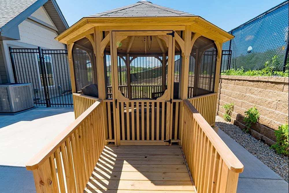 A wooden gazebo with a walkway leading to the entrance, surrounded by fencing on a sunny day.
