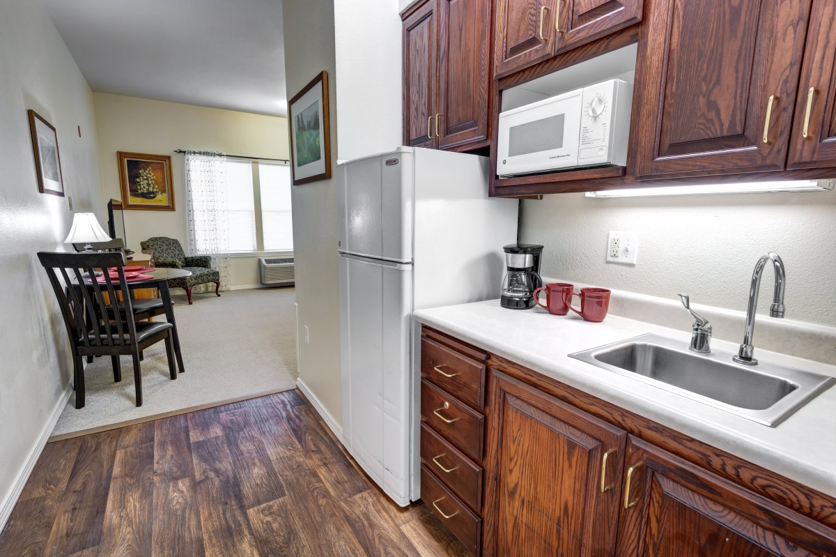A compact kitchen area with wooden cabinets, featuring a microwave, refrigerator, sink, and coffee maker. The adjacent dining area is visible in the background.