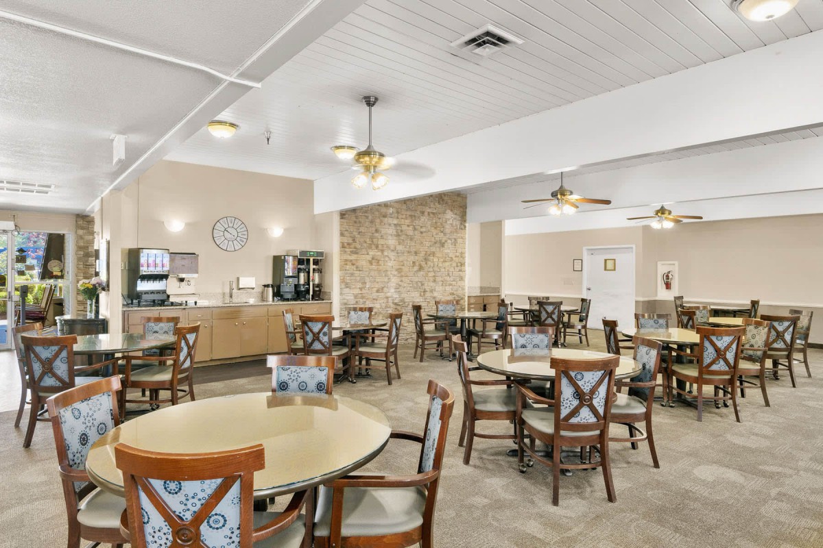 A spacious dining area with multiple round tables and chairs arranged neatly under ceiling fans, featuring a coffee station in the corner.