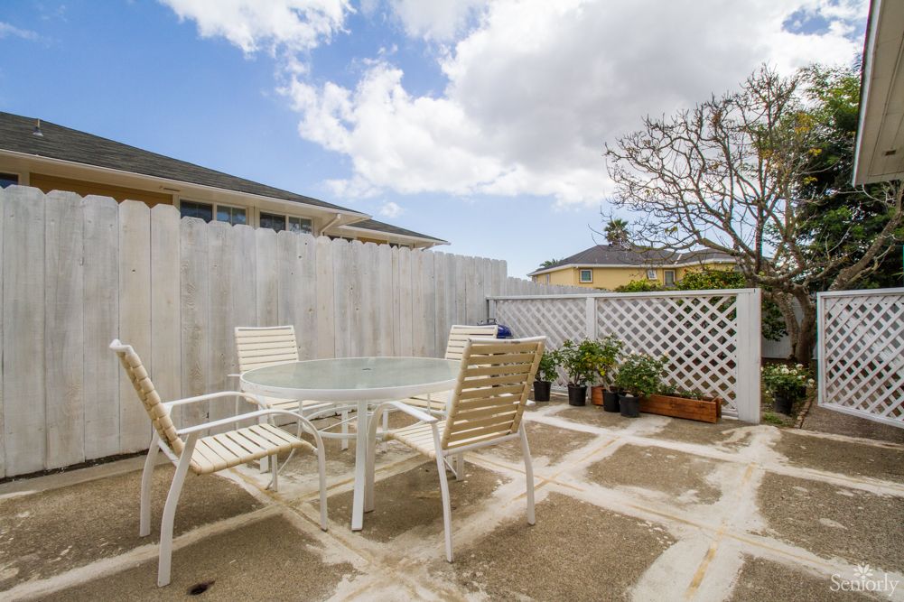A cozy patio with a glass table surrounded by chairs, enclosed by a fence with potted plants under a cloudy sky.