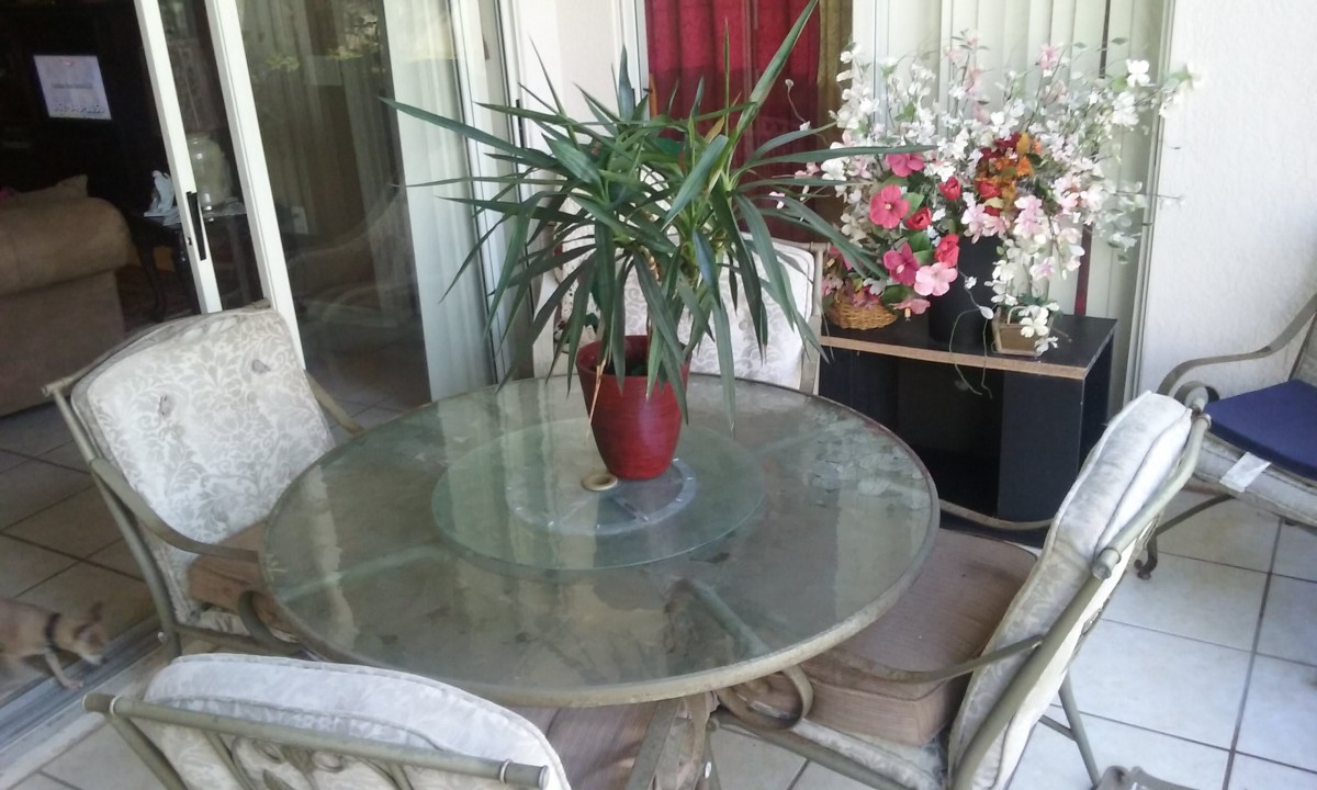 A cozy patio setting featuring a glass table with a potted plant centerpiece, surrounded by chairs and decorative flowers.