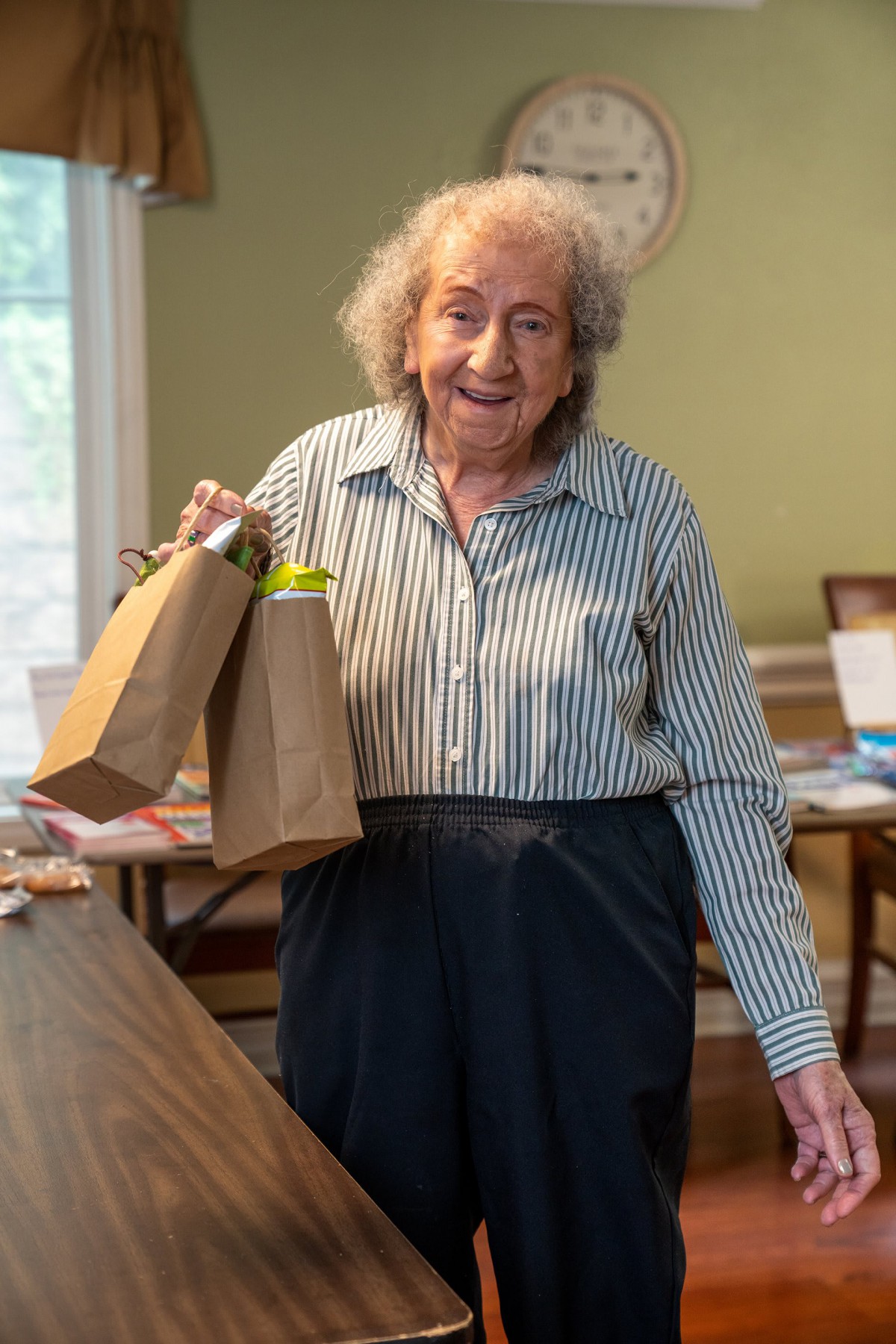 An elderly woman smiling and holding gift bags indoors.