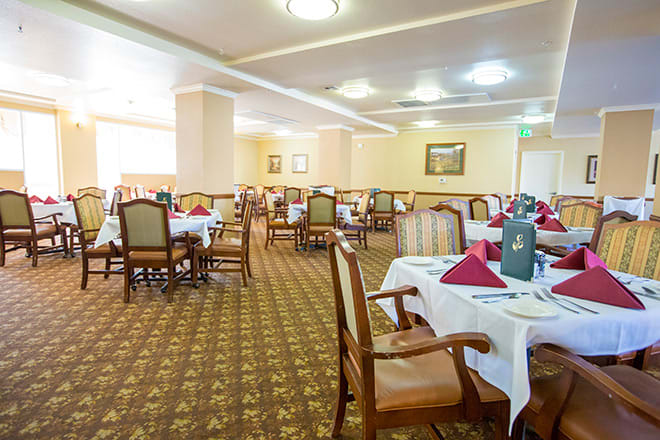 A spacious dining room with neatly arranged tables and chairs, featuring elegant table settings and carpeted flooring.