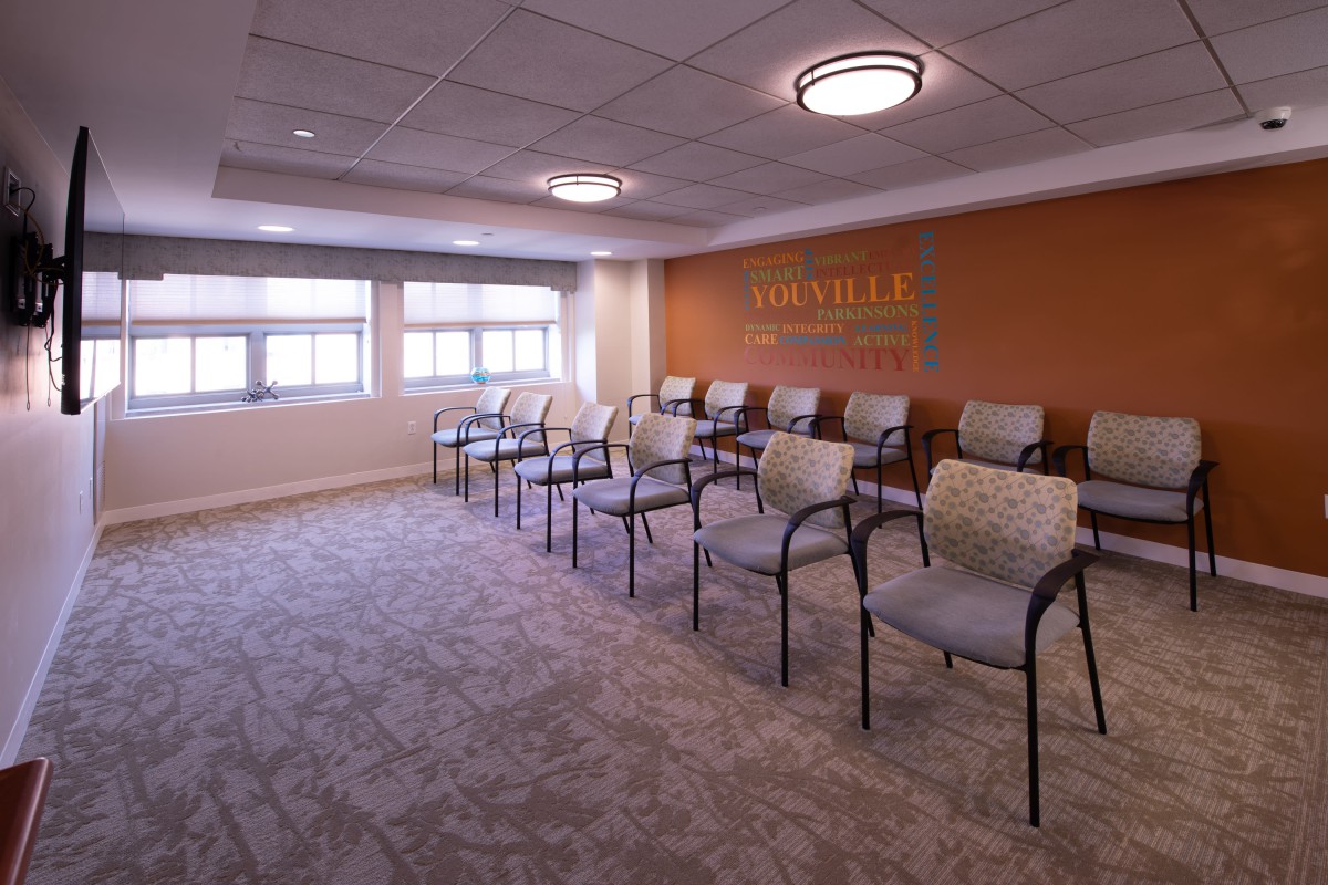 An indoor waiting room with several chairs arranged in rows, featuring windows and carpeted flooring.