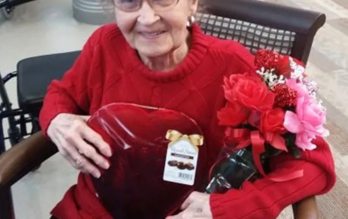 An elderly woman wearing a red sweater holding a gift box and a bouquet of flowers.