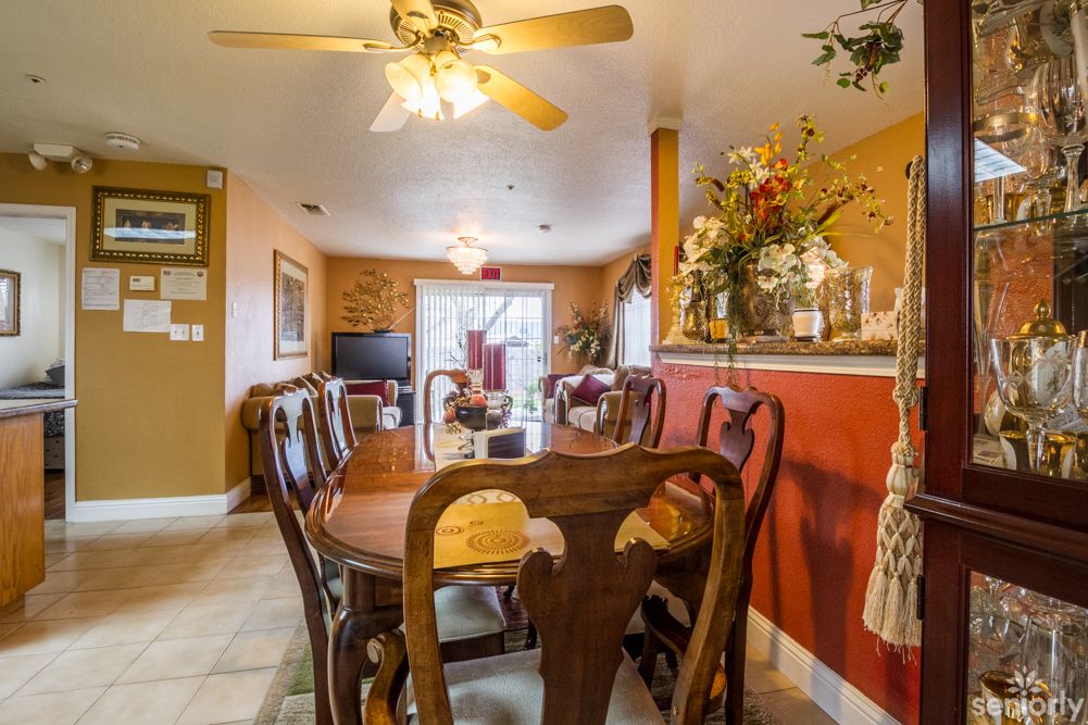 A cozy dining area with a wooden table and chairs, ceiling fan, and decorative flowers, leading into a living room.